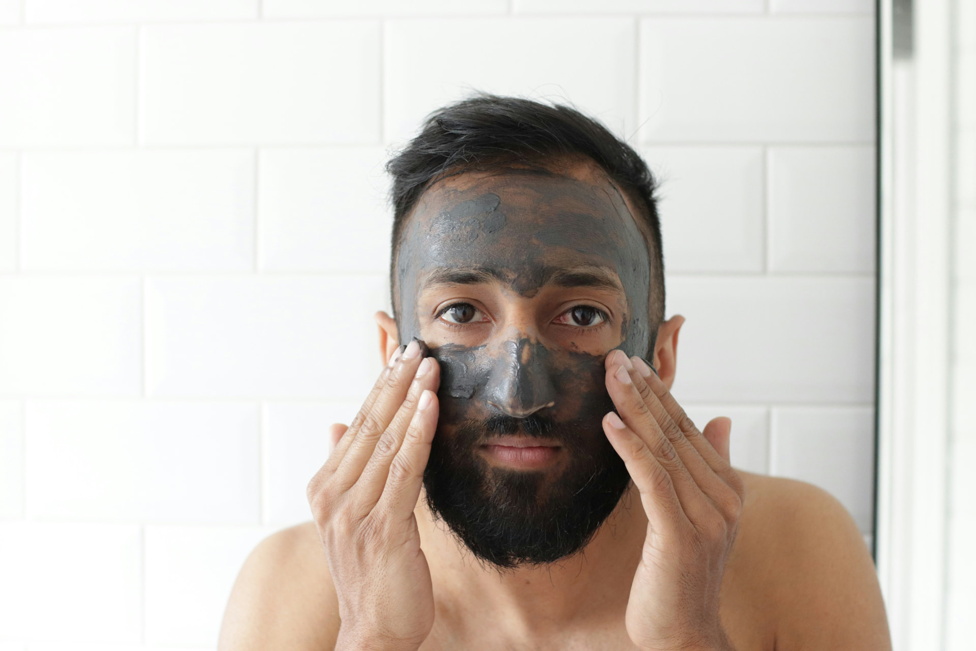 Uomo con barba applica una maschera viso all'argilla davanti a parete bianca