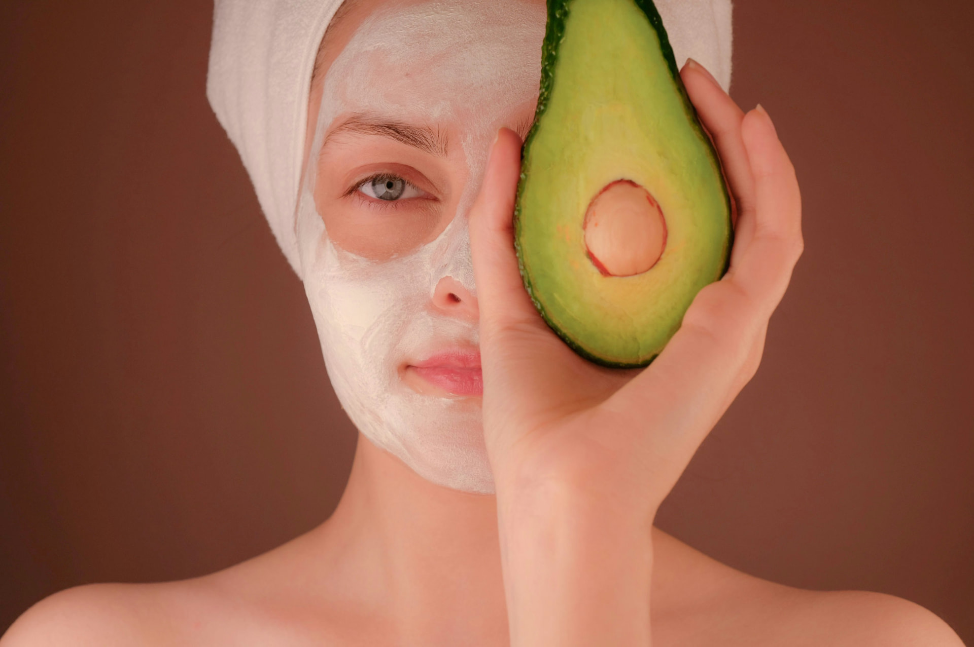 Donna con maschera viso in crema bianca e asciugamano tiene mezzo avocado davanti all'occhio