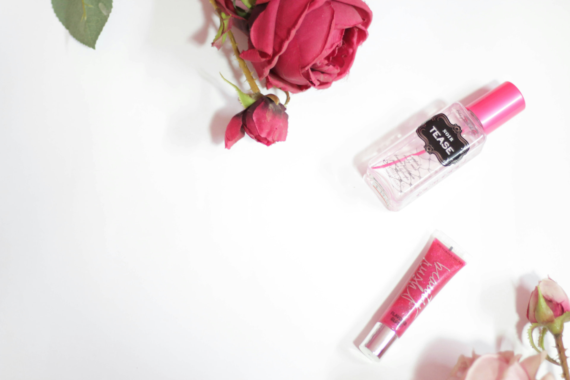 Body mist profumato e lucidalabbra rosa accanto a rose rosse su sfondo bianco